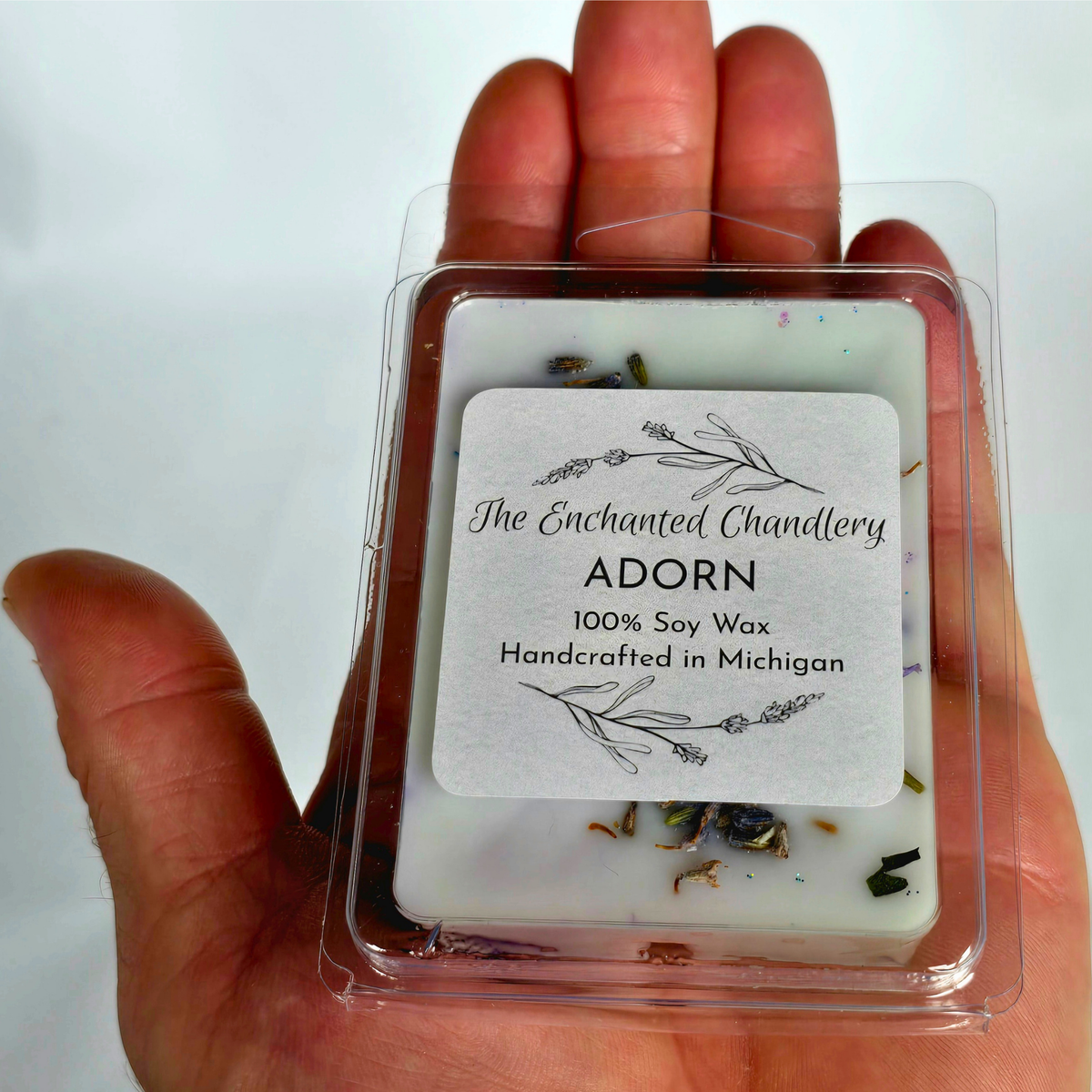 A hand holding a clear plastic clamshell package containing a square soy wax melt labeled 'The Enchanted Chandlery ADORN 100% Soy Wax Handcrafted in Michigan' with dried lavender and small botanical pieces visible inside.