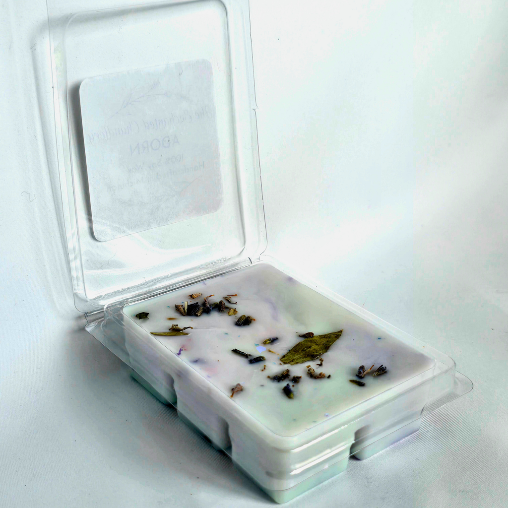 A clear plastic clamshell container holding a pale blue wax melt with scattered dried botanical pieces, shown open on a plain white background.