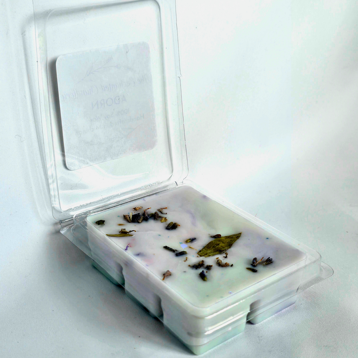 A clear plastic clamshell container holding a pale blue wax melt with scattered dried botanical pieces, shown open on a plain white background.