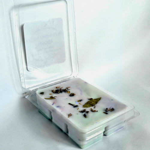 A clear plastic clamshell container holding a pale blue wax melt with scattered dried botanical pieces, shown open on a plain white background.