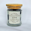 A glass jar candle with a light wood lid, filled with dark grey soy wax and dark charcoal wax on top. The front features a white label with black text that reads 'The Enchanted Chandlery EUXINE 100% Soy Wax Handcrafted in Michigan'.