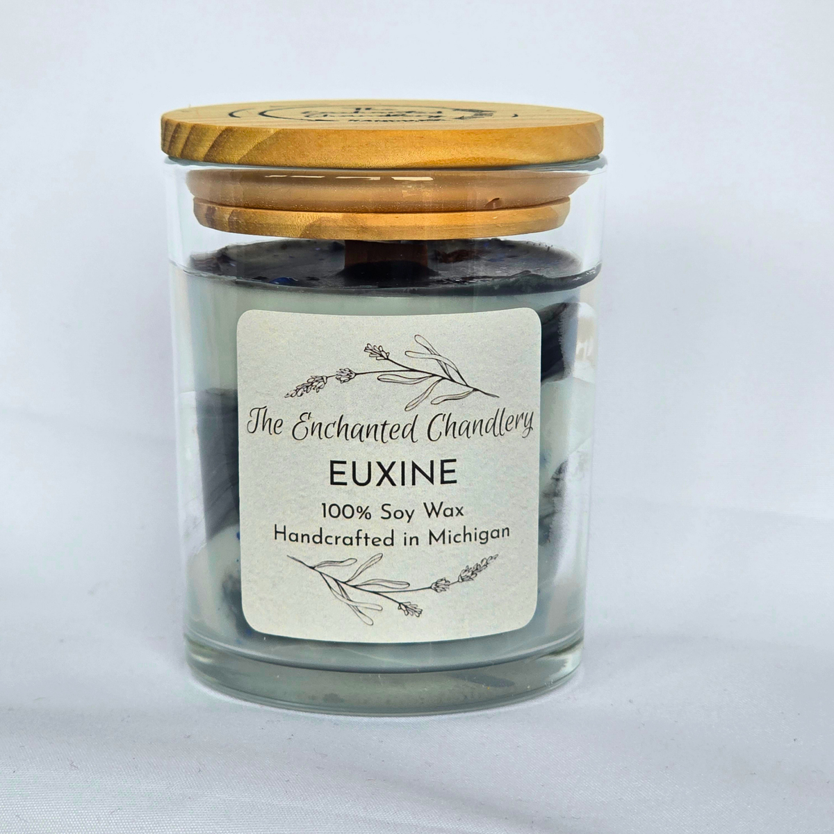 A glass jar candle with a light wood lid, filled with dark grey soy wax and dark charcoal wax on top. The front features a white label with black text that reads 'The Enchanted Chandlery EUXINE 100% Soy Wax Handcrafted in Michigan'.