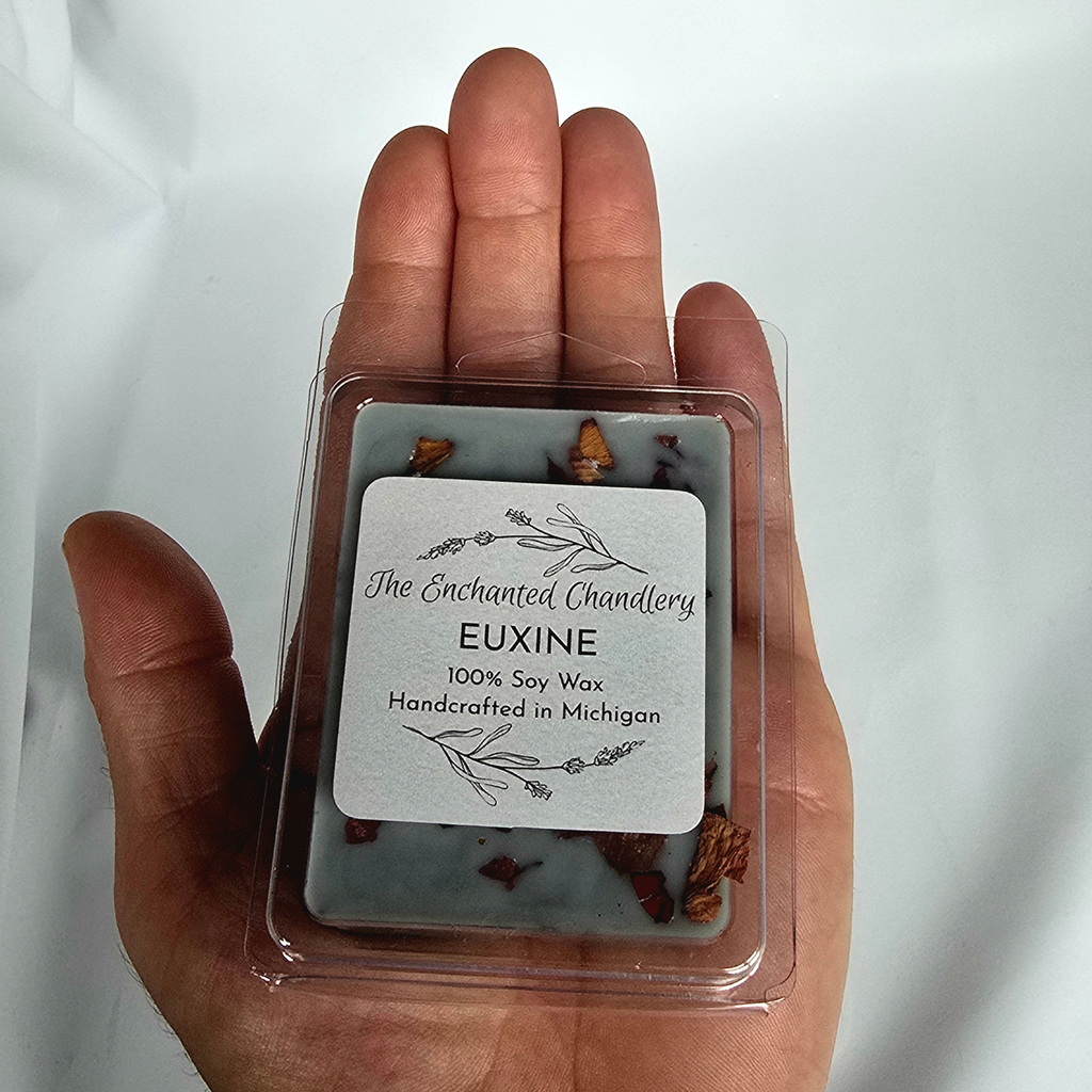 A hand holding a square clear plastic clamshell package of soy wax melts. The wax is a muted grey color with dried botanical pieces embedded, and the front label reads 'The Enchanted Chandlery EUXINE 100% Soy Wax Handcrafted in Michigan'.