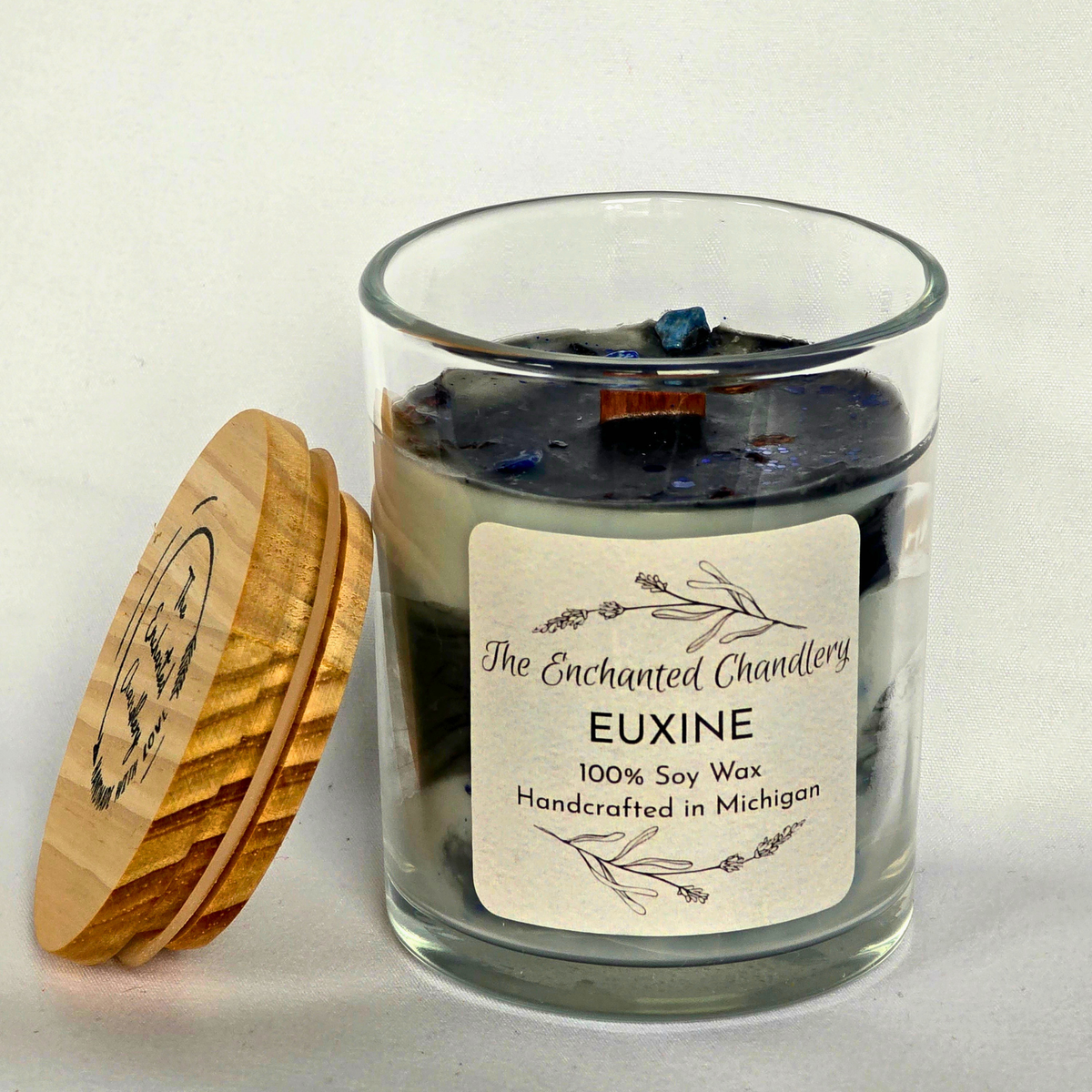 A glass jar candle labeled 'The Enchanted Chandlery EUXINE 100% Soy Wax' with a cream label and a wooden lid leaning against it on a light neutral background.
