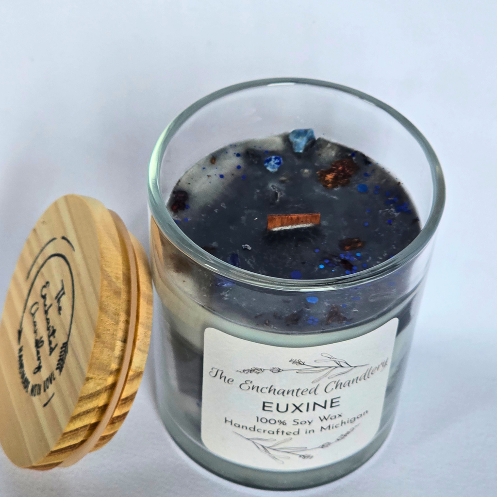 A glass jar candle labeled 'The Enchanted Chandlery' and 'EUXINE' with a white label, filled with dark wax and embedded with blue and copper glitter. A light wood lid with a circular logo sits beside the jar on a white background.
