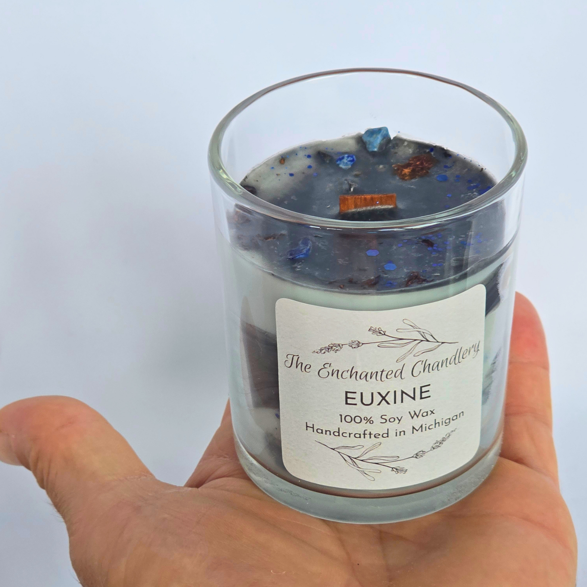 A hand holding a clear glass jar candle labeled 'The Enchanted Chandlery EUXINE 100% Soy Wax Handcrafted in Michigan'. The candle has a dark wax body with dark charcoal gray soot-like top layers containing blue, shown against a plain light background.