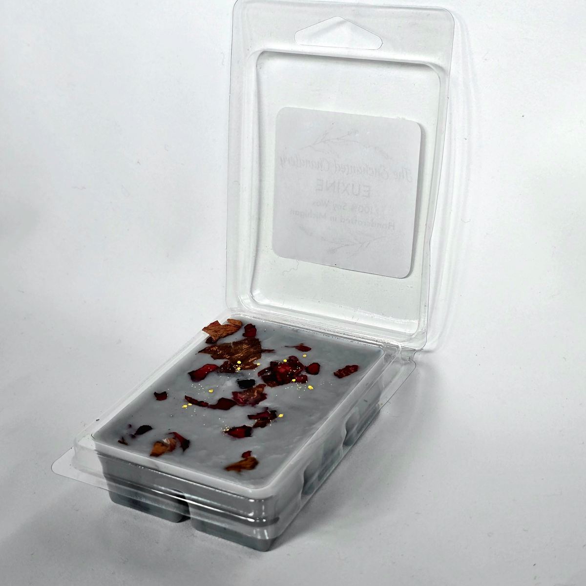 A clear plastic clamshell package containing a gray wax melt or wax melt block with scattered dried botanical pieces on top, photographed on a plain white background.