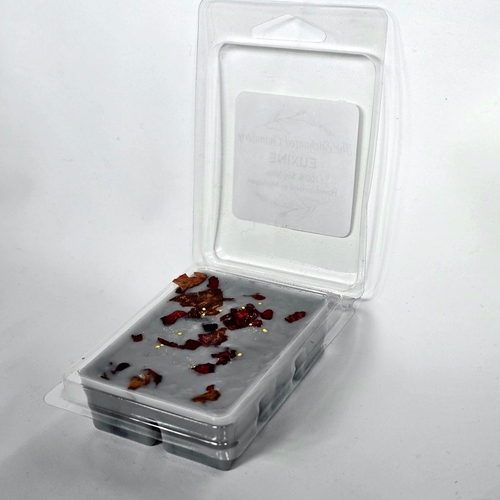 A clear plastic clamshell package containing a gray wax melt or wax melt block with scattered dried botanical pieces on top, photographed on a plain white background.