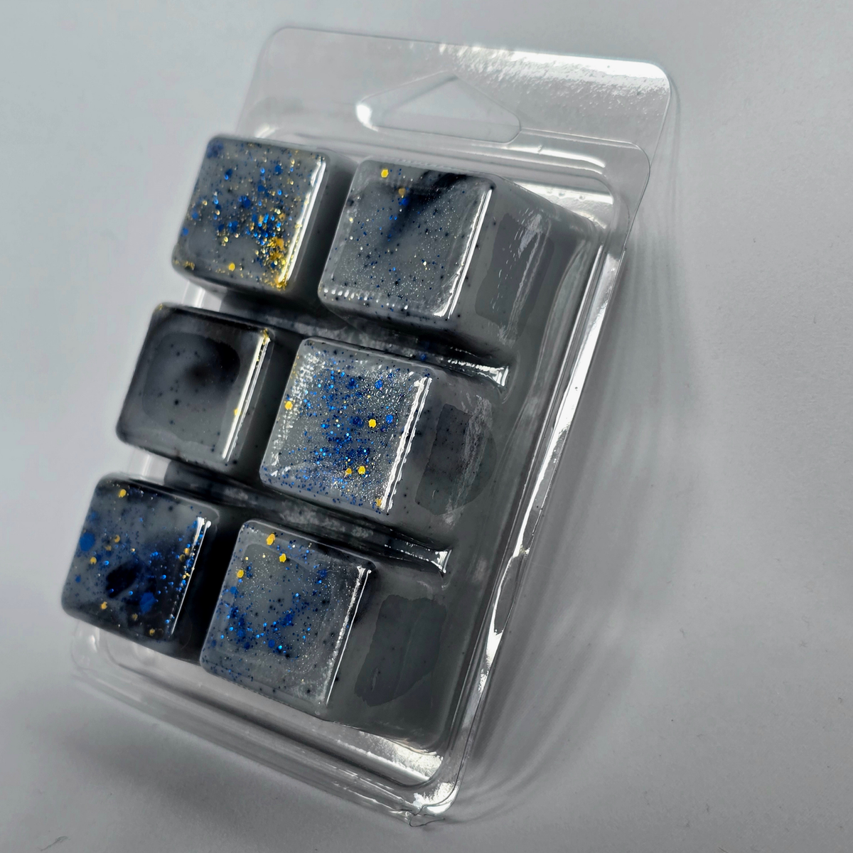 Six square wax melts in a clear plastic container, featuring a dark gray and black space-themed design with blue and gold glitter speckles.