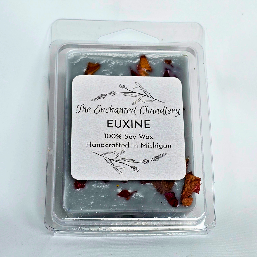 A clear plastic clamshell package of soy wax melts with a white label reading 'The Enchanted Chandlery EUXINE 100% Soy Wax Handcrafted in Michigan.' The wax is a pale grayish tone with scattered dried botanical pieces in muted red and  burgundy tones visible around the edges.
