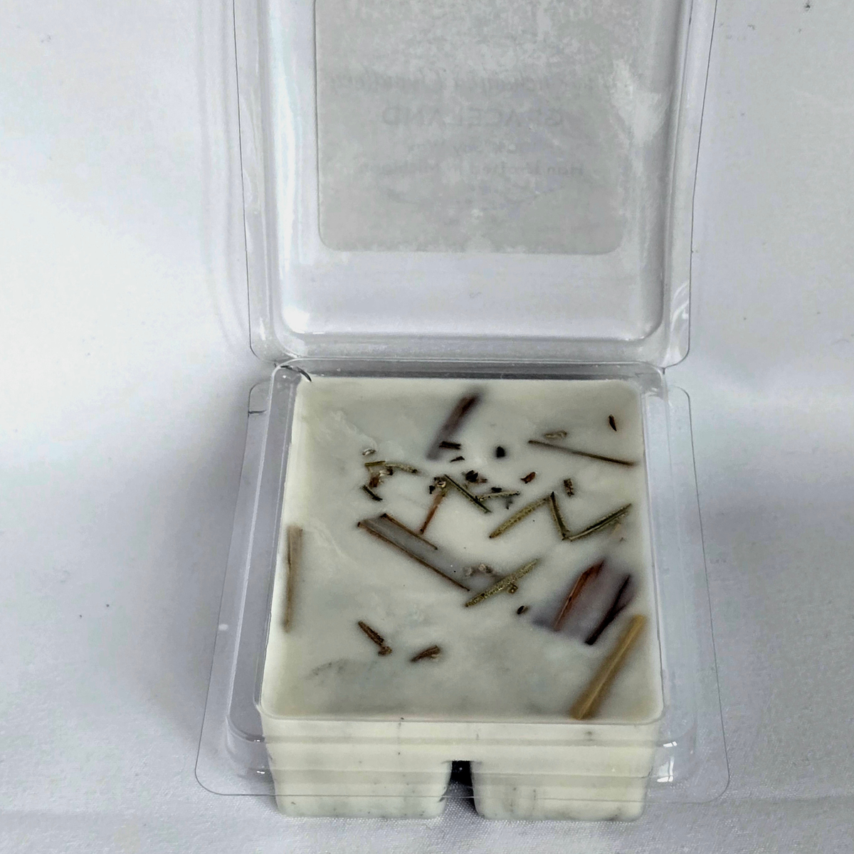 A square white wax melt with dried botanical inclusions, sealed in a clear plastic clamshell container with the lid open, photographed on a white background.