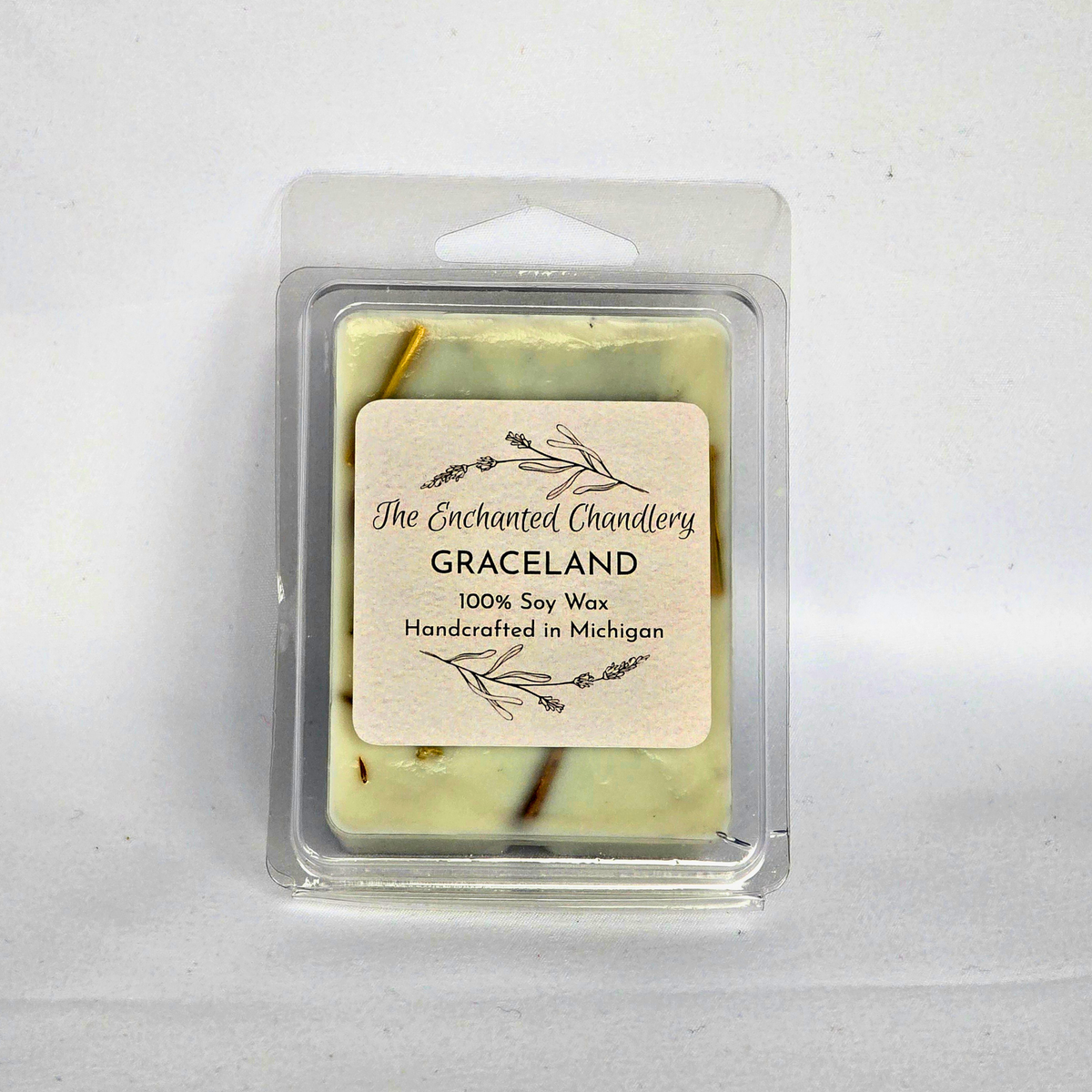 A packaged bar of soy wax melts from The Enchanted Chandlery, specifically the 'Graceland' scent. The wax is pale yellow with small dark brown botanical inclusions, housed in a clear plastic clamshell container with a cream-colored paper label.