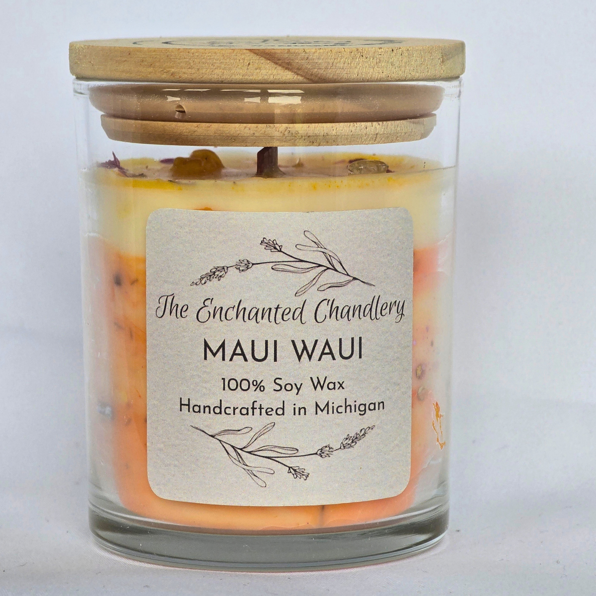 A glass jar candle with a light wood lid, filled with layered yellow and orange wax. A cream-colored paper label on the front reads 'The Enchanted Chandlery', 'MAUI WAUI', and '100% Soy Wax Handcrafted in Michigan'.