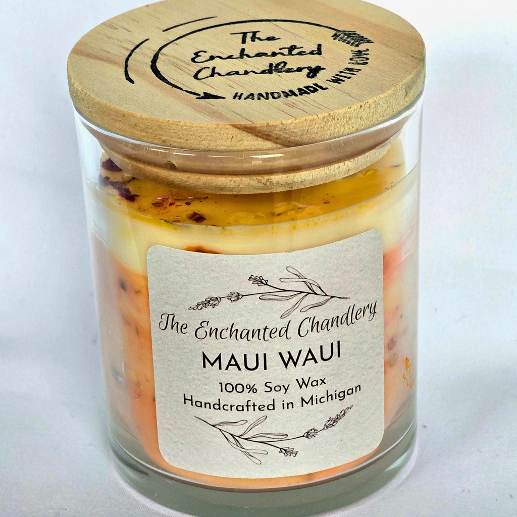 A glass jar candle with a light wood lid and a cream label reading 'The Enchanted Chandlery MAUI WAUI 100% Soy Wax Handcrafted in Michigan.' The candle wax inside shows layered peach, orange, and pale yellow tones with small dried botanical pieces near the top.