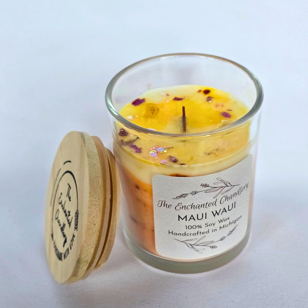 A glass jar candle with a light wood lid leaning beside it on a white background. The candle wax is pale yellow with scattered dried flower petals, and the front label reads 'The Enchanted Chandlery' and 'MAUI WAUI'.