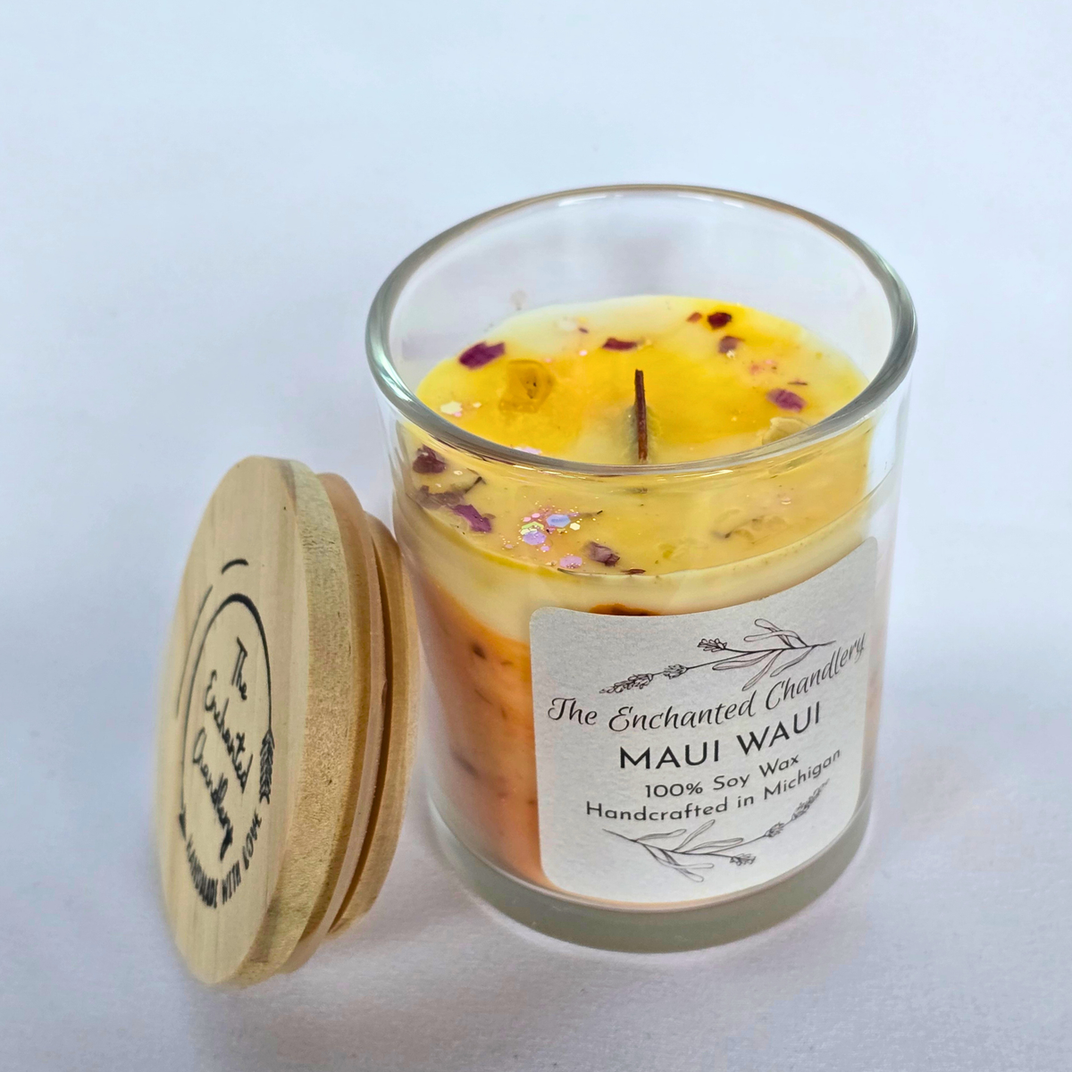 A glass jar candle with a light wood lid leaning beside it on a white background. The candle wax is pale yellow with scattered dried flower petals, and the front label reads 'The Enchanted Chandlery' and 'MAUI WAUI'.