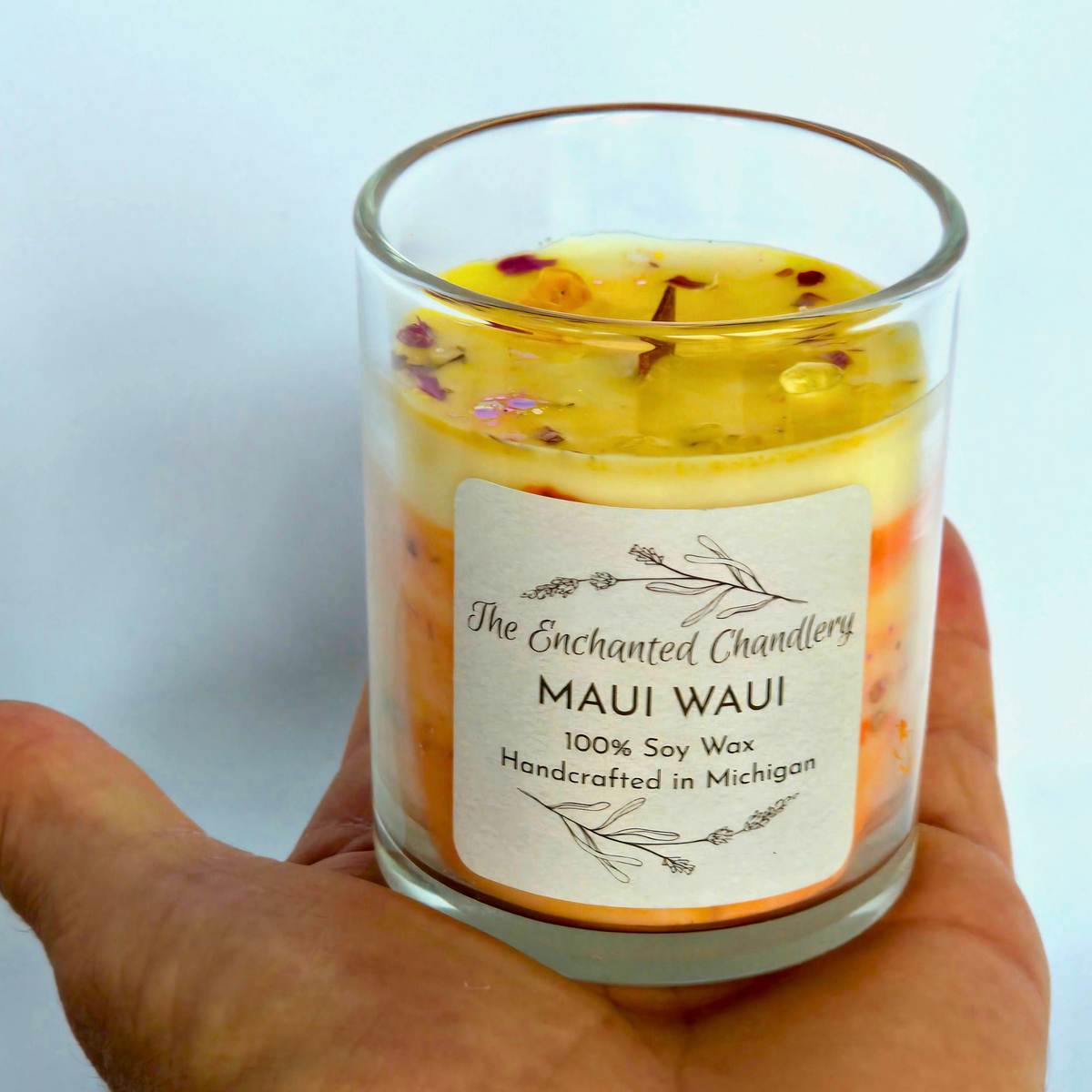 A hand holding a clear glass jar candle labeled 'The Enchanted Chandlery Maui Waui' with layered yellow, orange, and white wax and dried flower petals visible in the top.
