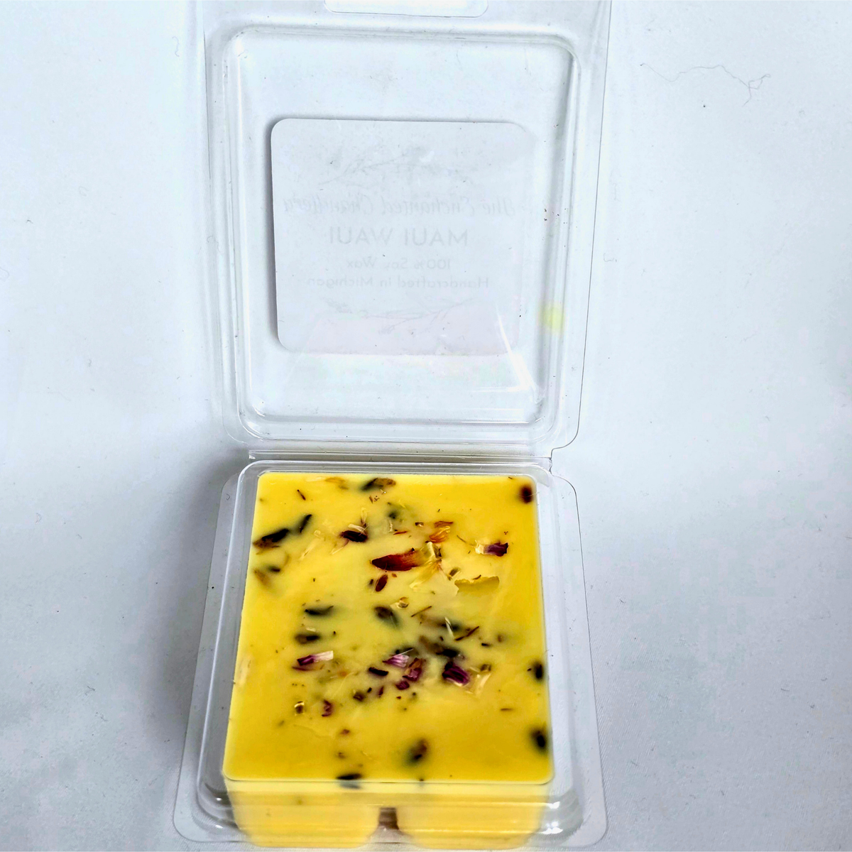 A square yellow wax melt in a clear plastic clamshell container, shown open against a plain light background. The wax has pink flecks and small dried botanical pieces on top.