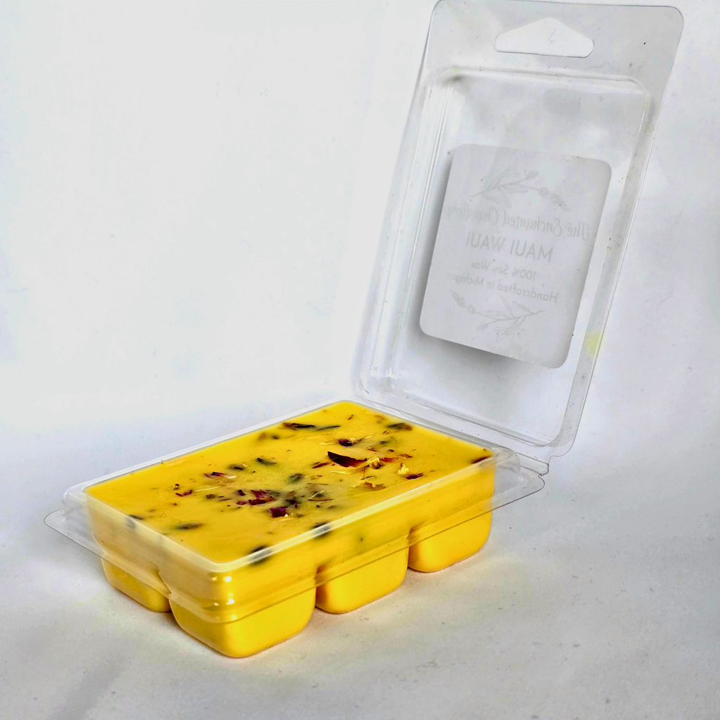 A yellow wax melt in a clear plastic clamshell container with an open hinged lid, showing a white paper insert card. The wax melt contains visible dried botanical pieces, and the packaging is photographed on a plain white background.