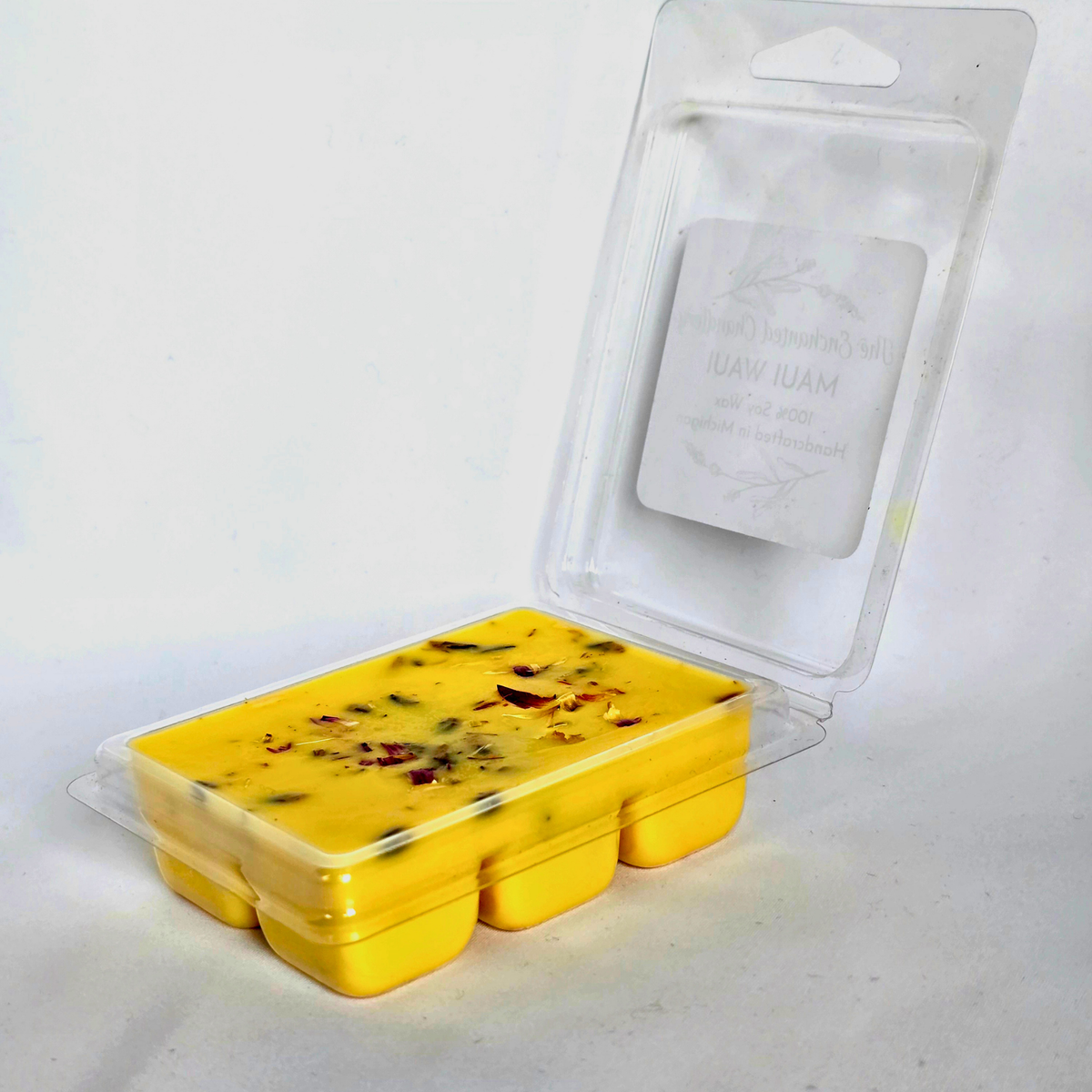 A yellow wax melt in a clear plastic clamshell container with an open hinged lid, showing a white paper insert card. The wax melt contains visible dried botanical pieces, and the packaging is photographed on a plain white background.