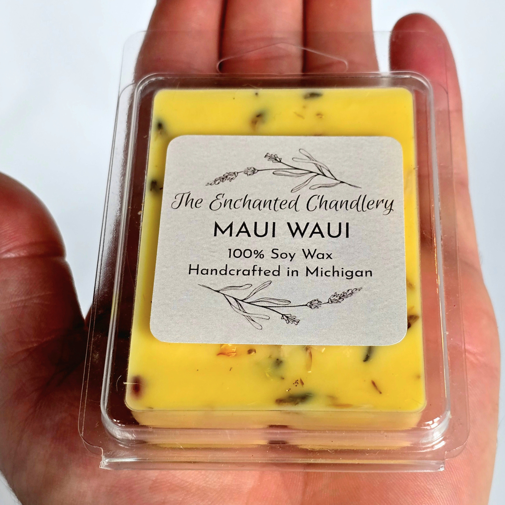 A hand holding a clear plastic clamshell package containing a yellow Maui Waui soy wax melt. The wax melt has small  pink and orange botanical flecks, and the label reads 'The Enchanted Chandlery MAUI WAUI 100% Soy Wax Handcrafted in Michigan'.