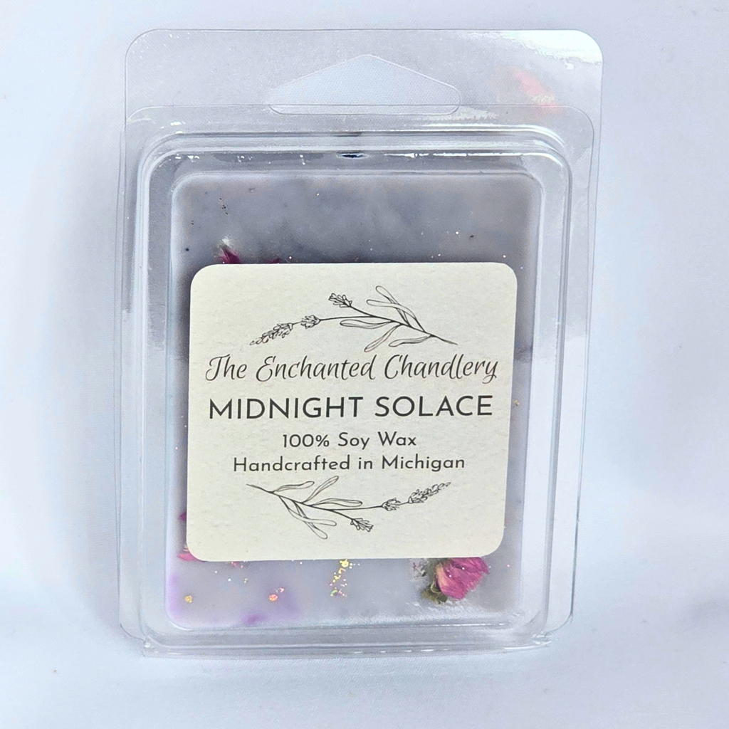 A clear plastic clamshell package containing soy wax melts with a cream label reading 'The Enchanted Chandlery MIDNIGHT SOLACE 100% Soy Wax Handcrafted in Michigan'. The wax appears smoky gray and dark purple with small pink and gold botanical accents visible near the bottom.