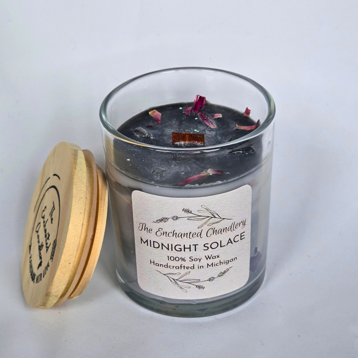A glass jar candle with dark gray wax and dried flower petals inside, shown with a light wood lid leaning against it on a plain light background. The front label reads 'The Enchanted Chandlery Midnight Solace 100% Soy Wax Handcrafted in Michigan.