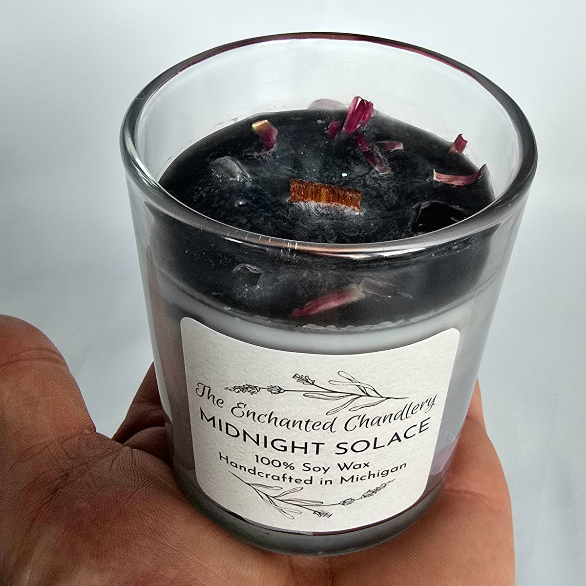 A hand holding a glass jar candle labeled 'Midnight Solace' by The Enchanted Chandlery. The candle has a white wax base with a dark charcoal/black top layer, embedded ice, and scattered dried rose petals.