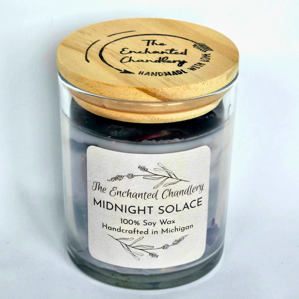 A glass jar candle with pale purple soy wax and a light wood lid engraved with 'The Enchanted Chandlery'. A white paper label on the front reads 'MIDNIGHT SOLACE' and '100% Soy Wax'.