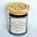 A glass jar candle with pale purple soy wax and a light wood lid engraved with 'The Enchanted Chandlery'. A white paper label on the front reads 'MIDNIGHT SOLACE' and '100% Soy Wax'.