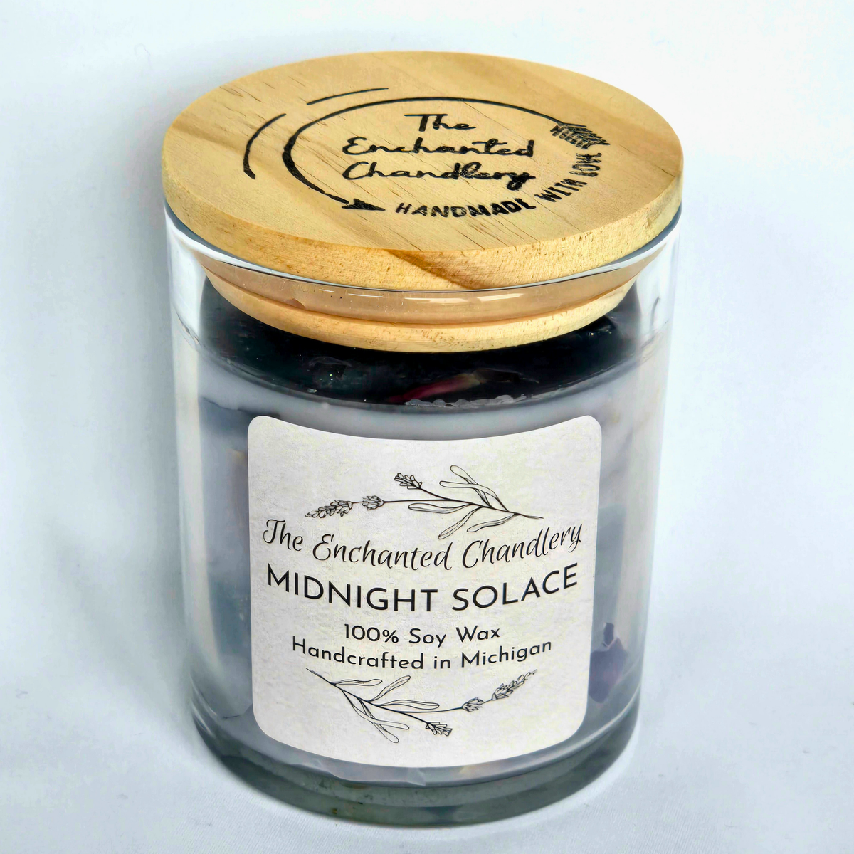 A glass jar candle with pale purple soy wax and a light wood lid engraved with 'The Enchanted Chandlery'. A white paper label on the front reads 'MIDNIGHT SOLACE' and '100% Soy Wax'.
