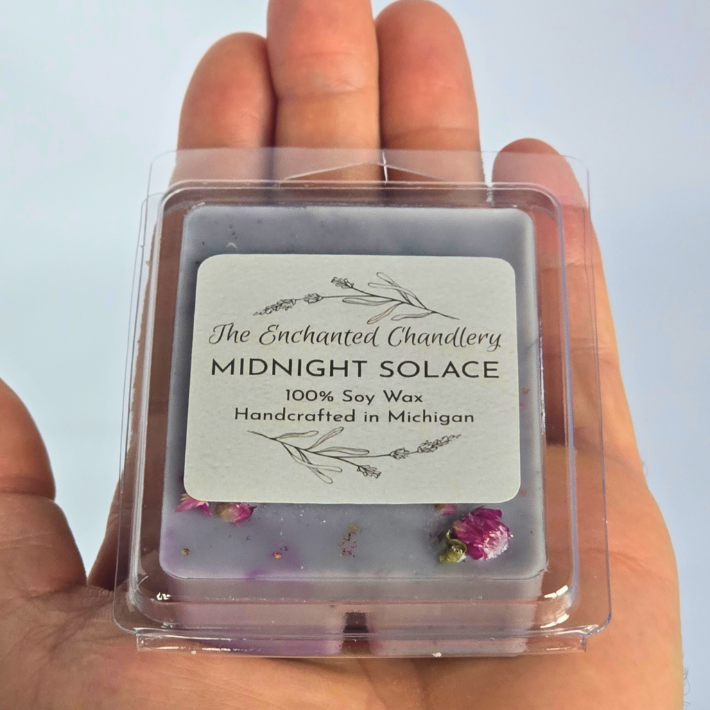 A hand holding a square clear plastic clamshell package of soy wax melts. The wax is a muted lavender-gray color with dried pink flowers and glitter, and a white label reading 'The Enchanted Chandlery MIDNIGHT SOLACE 100% Soy Wax Handcrafted in Michigan'.