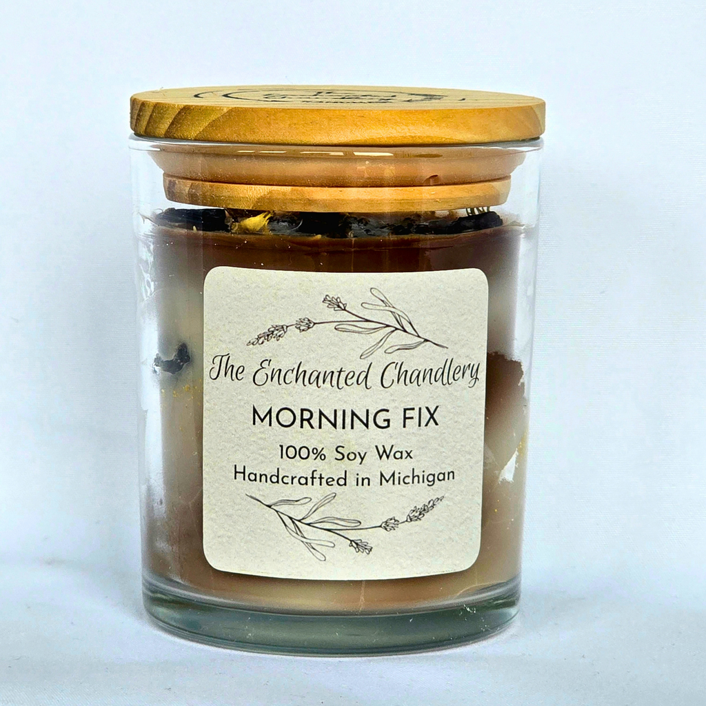 Clear glass jar candle filled with shades of brown wax, adorned with coffee beans and a light wood colored lid. Cream colored label with "The Enchanted Chandlery MORNING FIX"