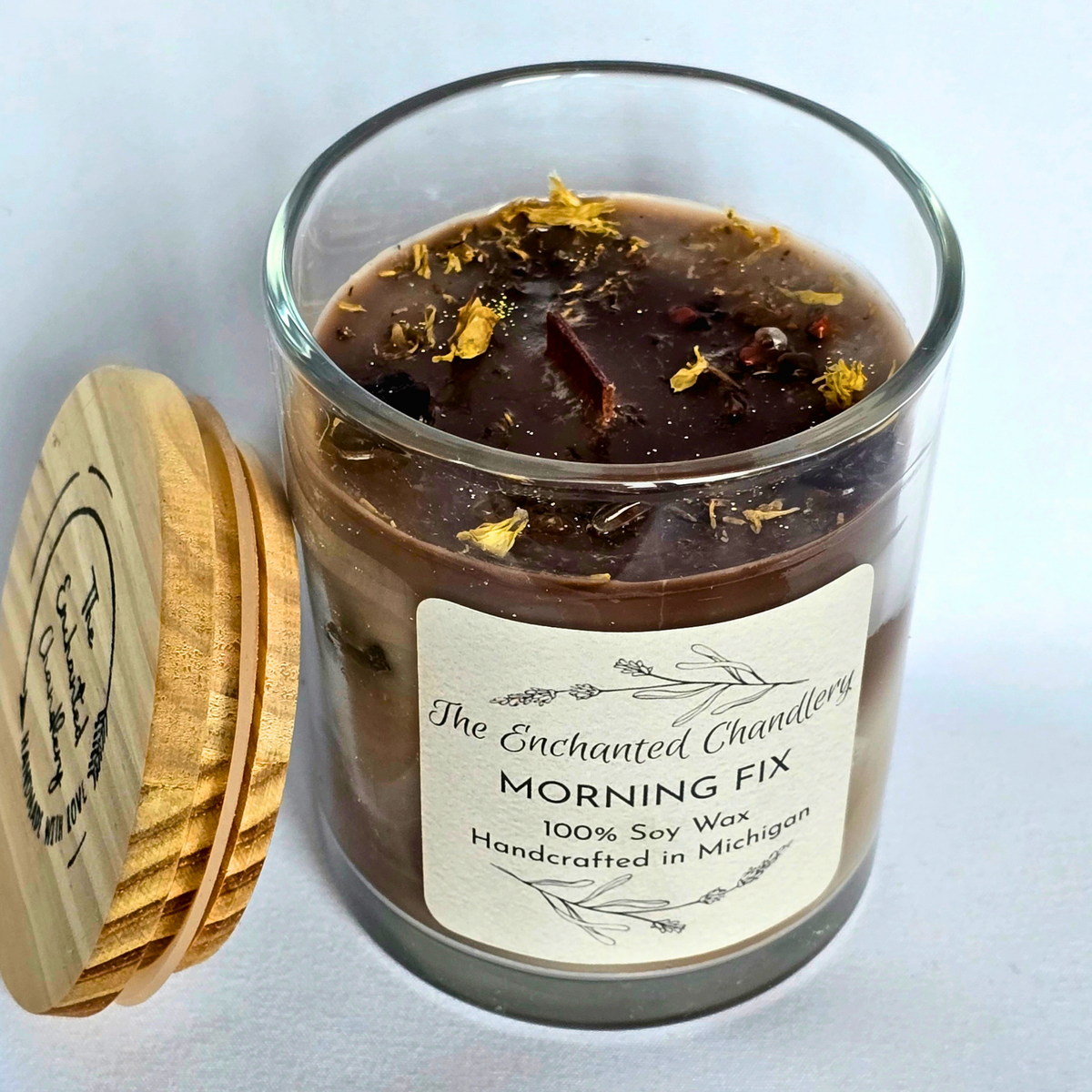 Clear glass jar candle filled with shades of brown wax, adorned with coffee beans and pale yellow colored botanicals. Cream colored label with "The Enchanted Chandlery MORNING FIX"