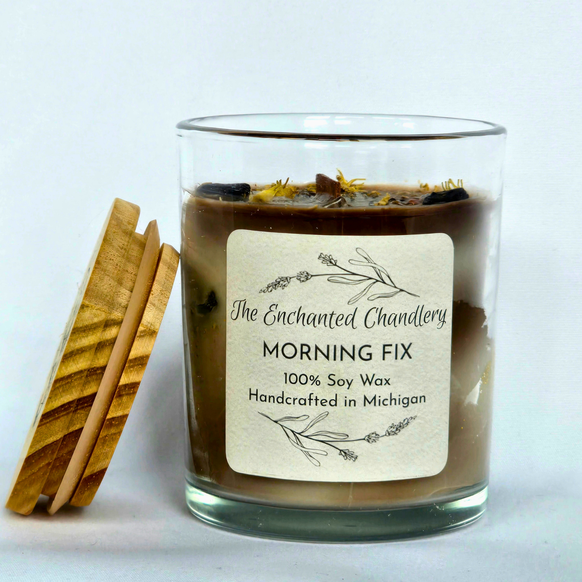 Clear glass jar candle filled with shades of brown wax, adorned with coffee beans and a light wood colored lid. Cream colored label with "The Enchanted Chandlery MORNING FIX"