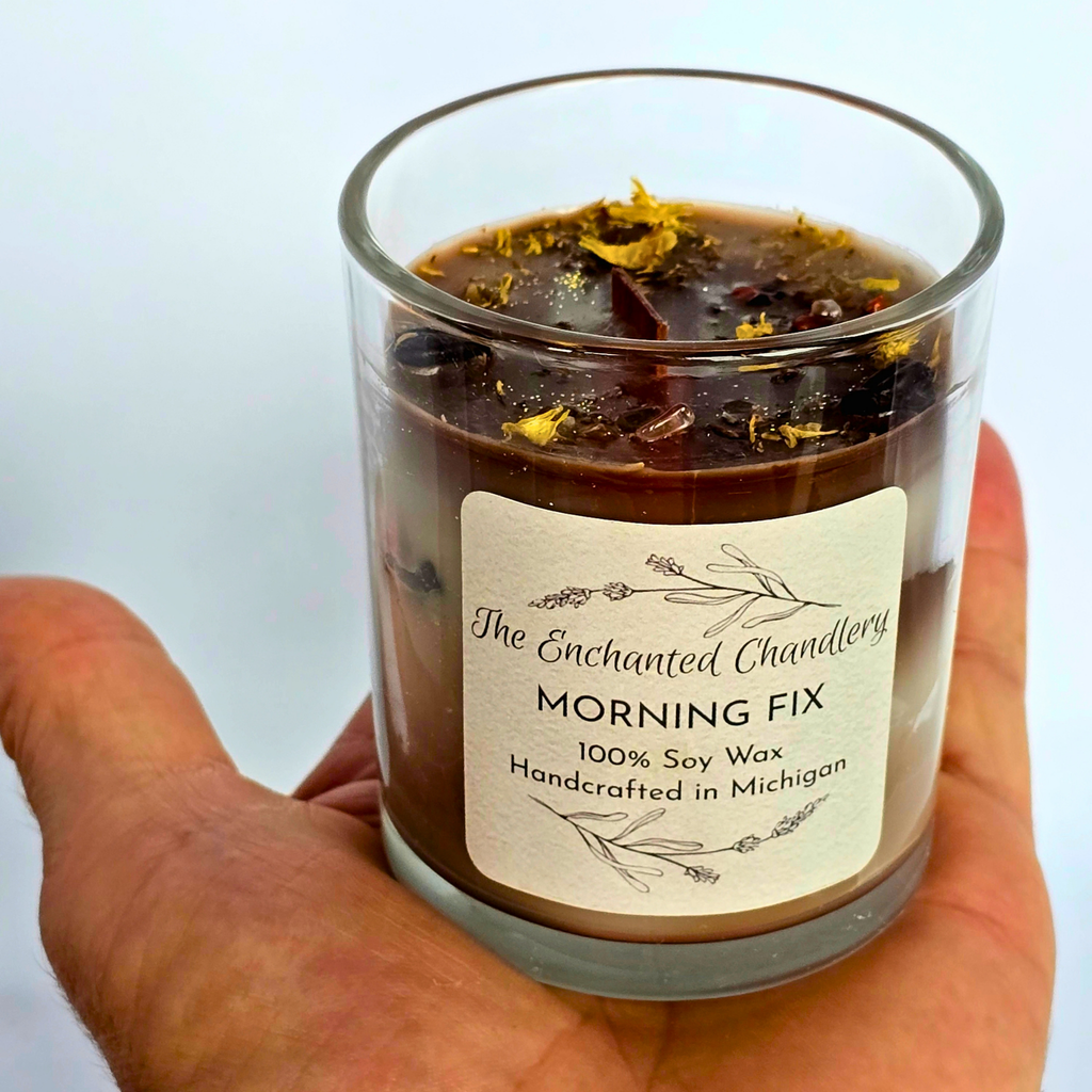 Hand holding a clear glass jar candle filled with shades of brown wax, adorned with coffee beans and a wood wick. Cream colored label with "The Enchanted Chandlery MORNING FIX"