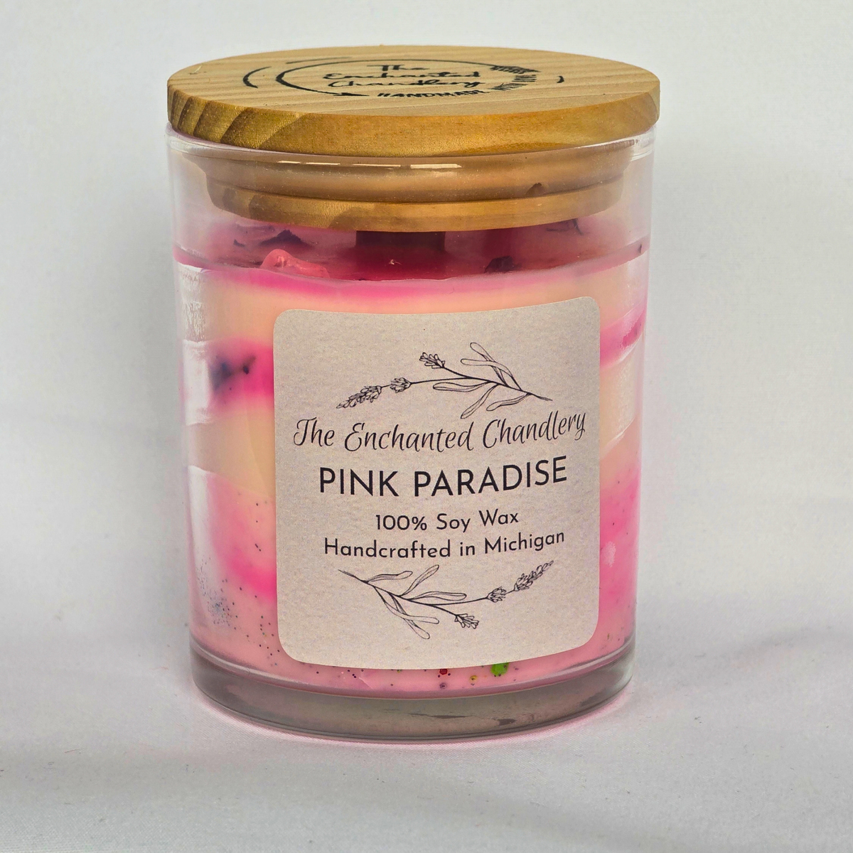 A glass jar candle with pale pink wax and dried botanical pieces on top, shown with a light wooden lid on it with a light gray background. The front label reads 'The Enchanted Chandlery Pink Paradise'