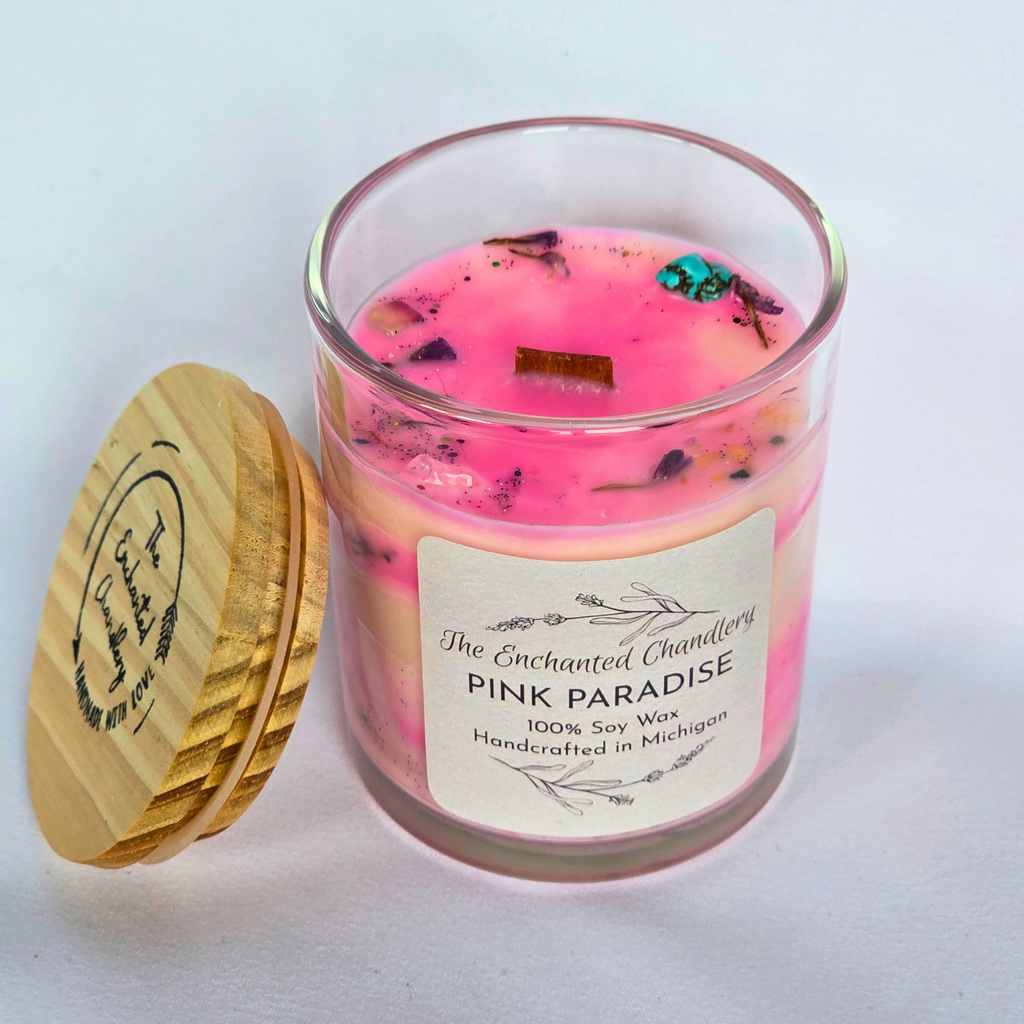 A glass jar candle with pale pink wax and dried botanical pieces on top, shown with a light wooden lid beside it on a light gray background. The front label reads 'The Enchanted Chandlery Pink Paradise'.
