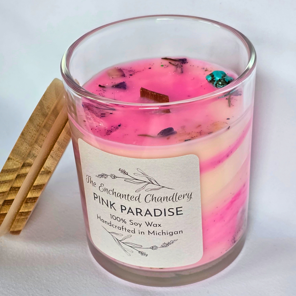 A glass jar candle with pale pink wax and dried botanical pieces on top, shown with a light wooden lid beside it on a light gray background. The front label reads 'The Enchanted Chandlery Pink Paradise'.