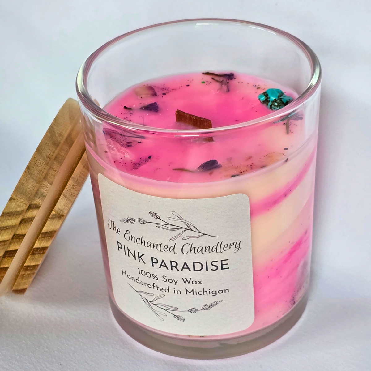 A glass jar candle with pale pink wax and dried botanical pieces on top, shown with a light wooden lid beside it on a light gray background. The front label reads 'The Enchanted Chandlery Pink Paradise'.