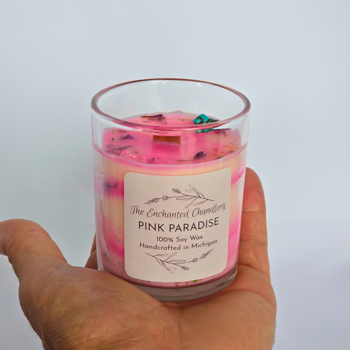 A hand holding a glass jar candle with pale pink wax and dried botanical pieces on top, shown with a light wooden lid beside it on a light gray background. The front label reads 'The Enchanted Chandlery Pink Paradise'.