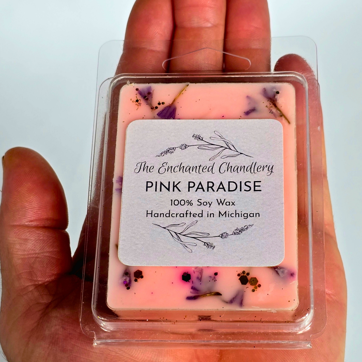 A hand holding a clear plastic clamshell package of soy wax melts. The wax is light pink with scattered purple flower petals and small dark specks, and the front label reads 'The Enchanted Chandlery PINK PARADISE 100% Soy Wax Handcrafted in Michigan'.