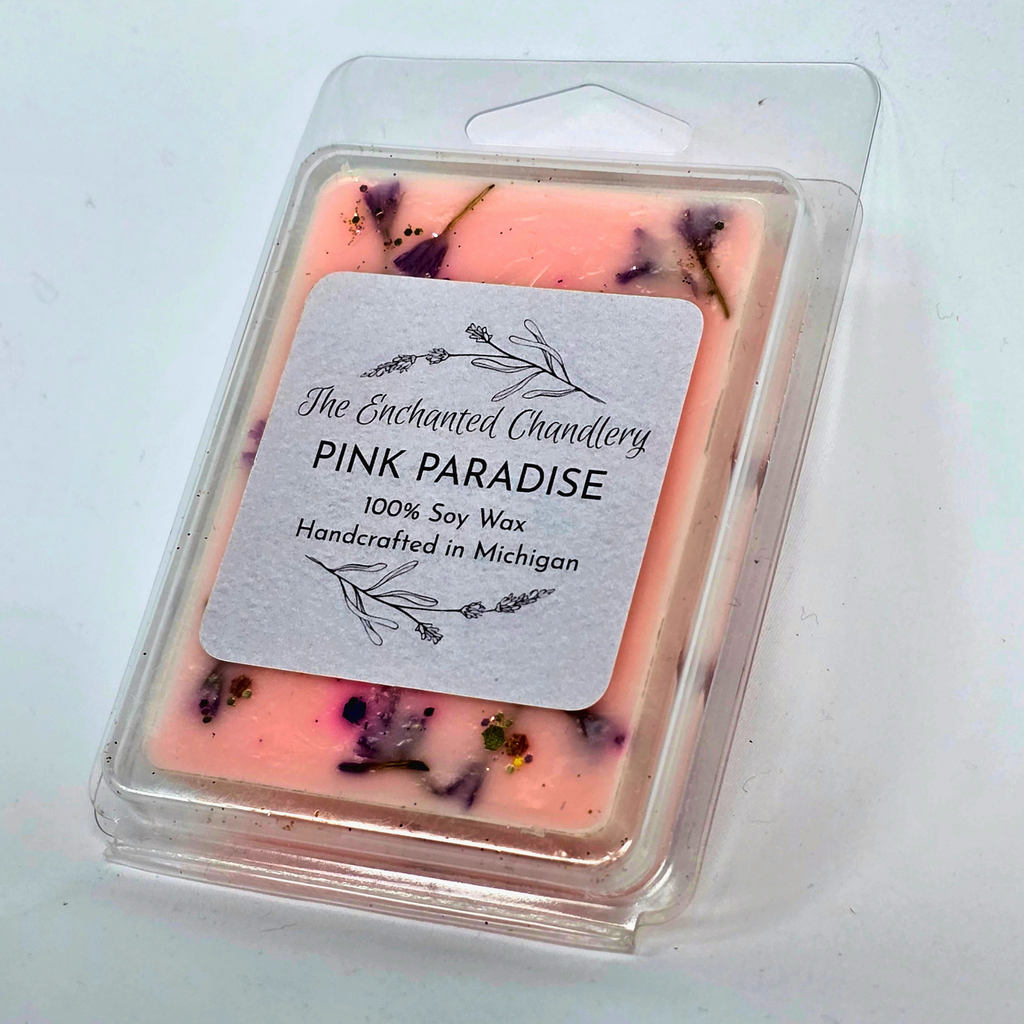 A packaged wax melt in a clear plastic clamshell container. The wax is light pink with embedded purple dried flower petals, and a white paper label on the front reads 'The Enchanted Chandlery PINK PARADISE 100% Soy Wax Handcrafted in Michigan'.