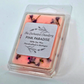 A packaged wax melt in a clear plastic clamshell container. The wax is light pink with embedded purple dried flower petals, and a white paper label on the front reads 'The Enchanted Chandlery PINK PARADISE 100% Soy Wax Handcrafted in Michigan'.