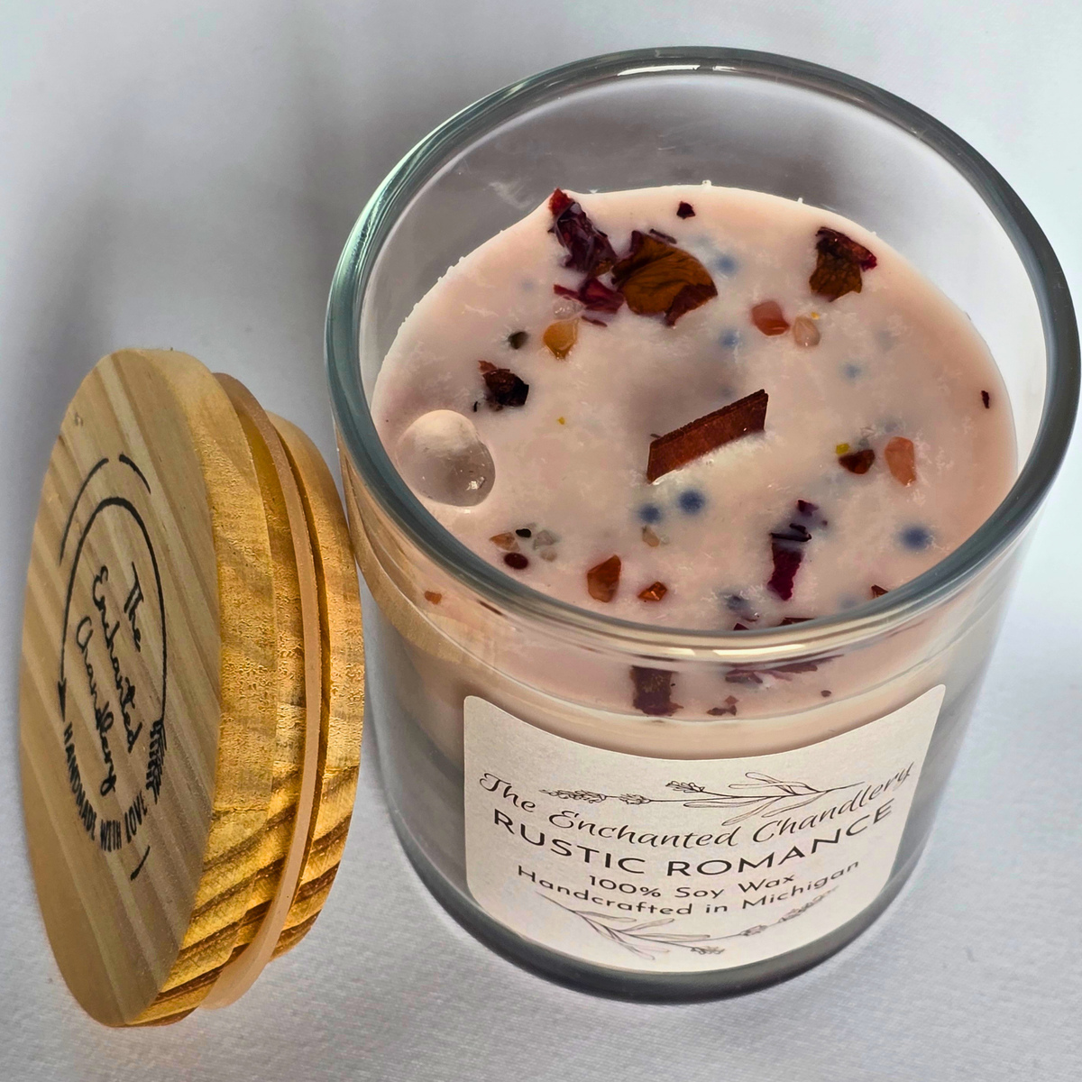 A glass jar candle with pale pink wax and scattered botanical flecks, shown next to its round wooden lid on a light neutral background. The front label reads 'The Enchanted Chambering Rustic Romance' and '100% Soy Wax Handcrafted in Michigan.