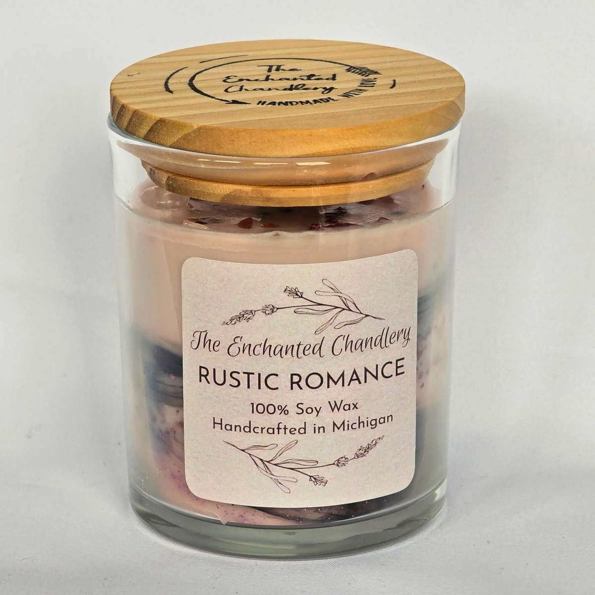 A glass jar candle with pale pink wax and dried botanical pieces on top, shown with a wooden lid on a light gray background. The front label reads 'The Enchanted Chandlery Rustic Romance'.