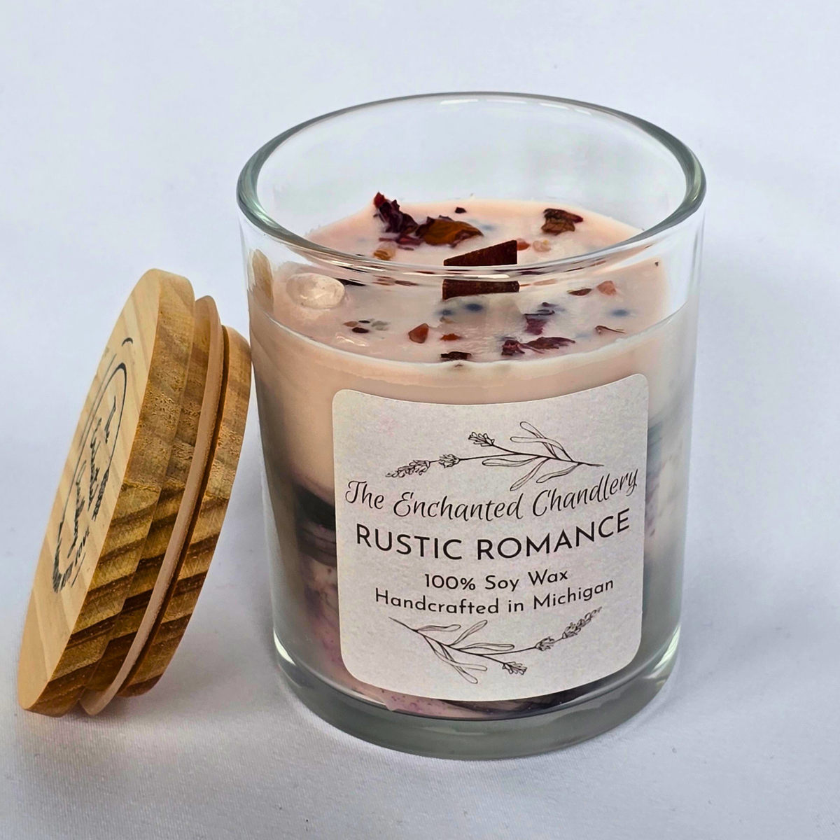 A glass jar candle with pale pink wax and dried botanical pieces on top, shown with a stack of wooden lids beside it on a light gray background. The front label reads 'The Enchanted Chandlery Rustic Romance'.