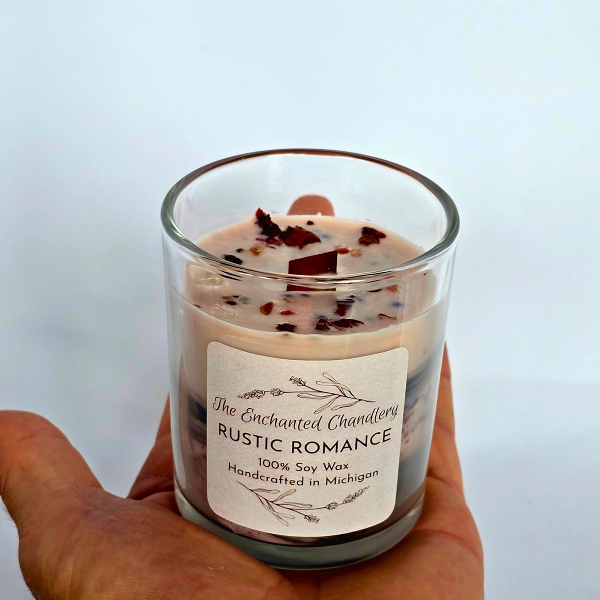 A hand holding a clear glass jar candle labeled 'The Enchanted Chandlery Rustic Romance' with cream-colored soy wax and dark red botanical pieces on top, against a plain light background.