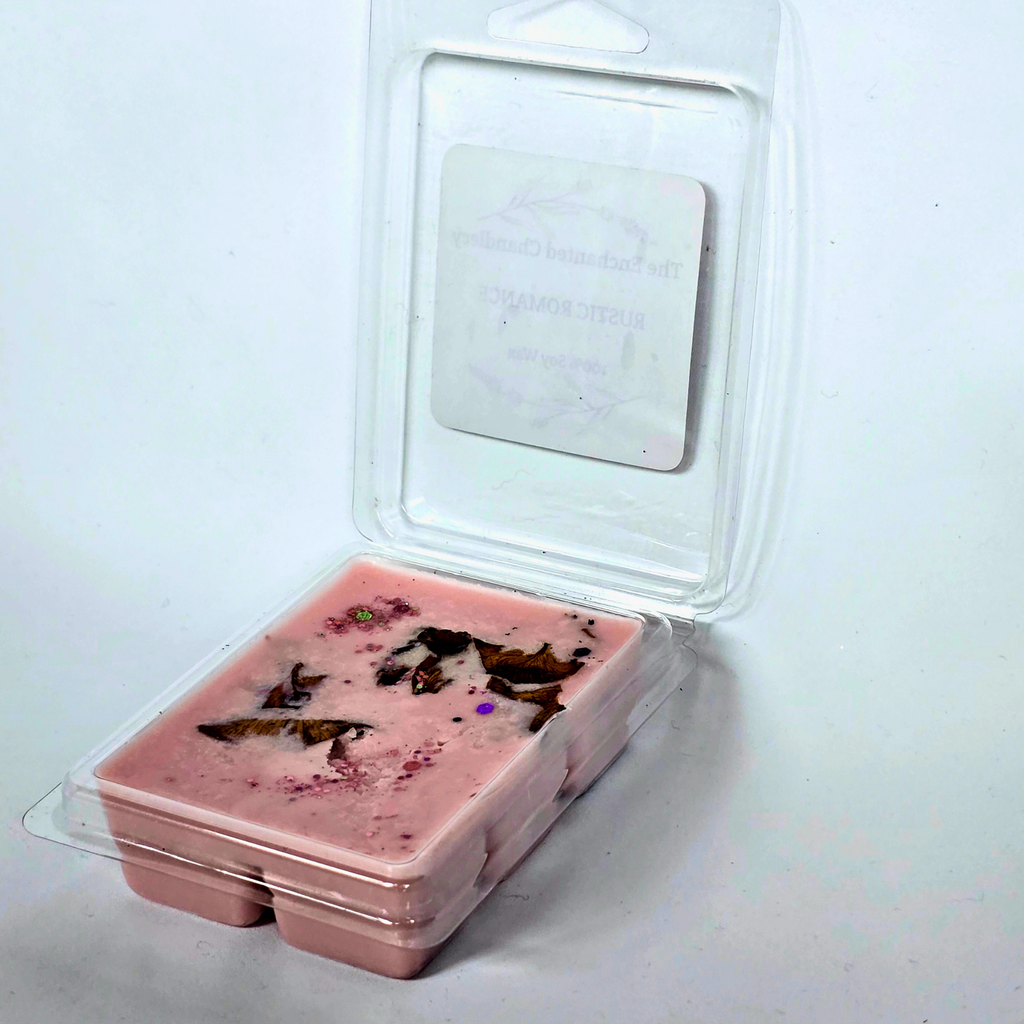 A pink wax melt in a clear plastic clamshell container with a hinged lid, photographed on a plain light background. The wax has red dried botanical pieces embedded in it, and a white label is visible inside the lid.