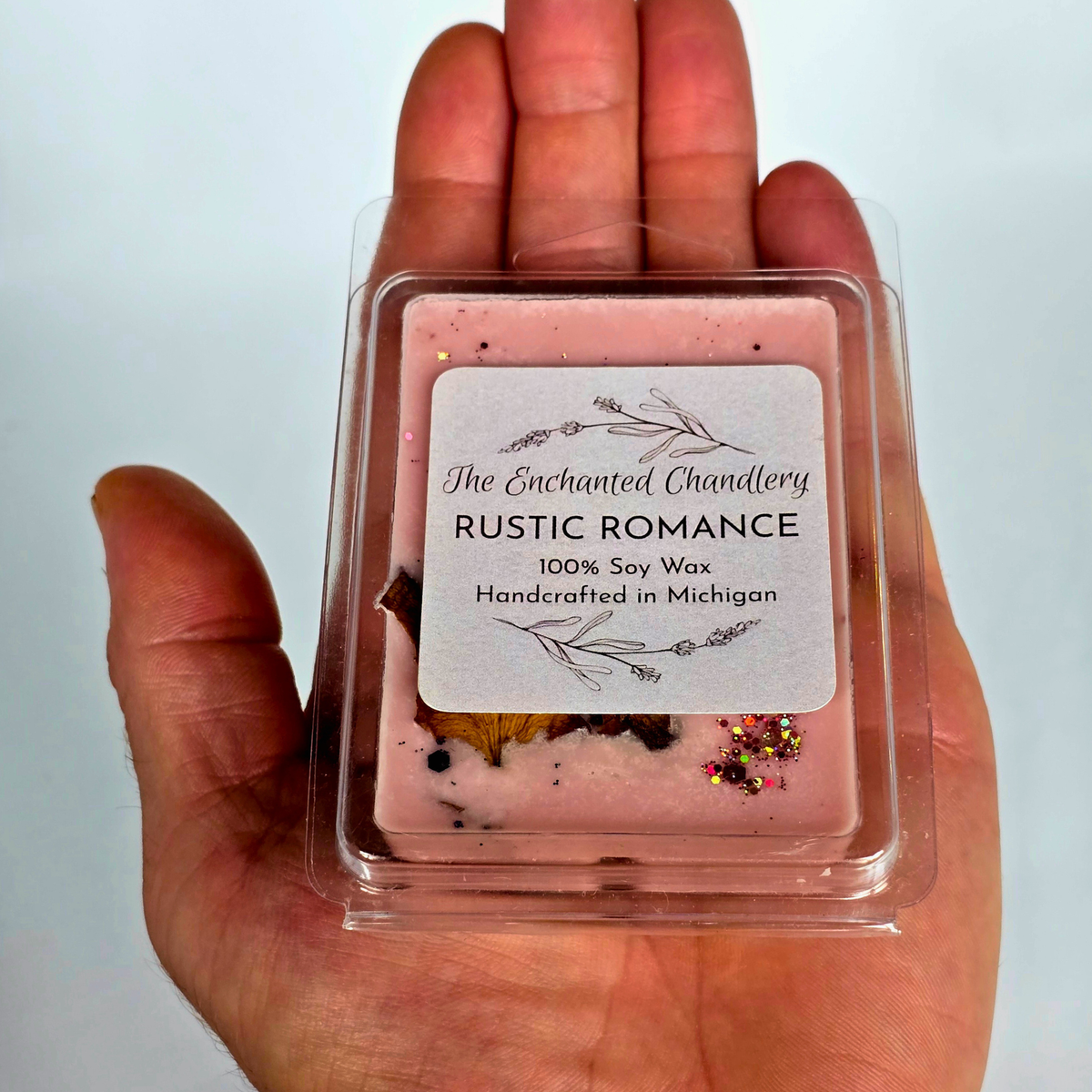 A hand holding a clear plastic clamshell container of pink soy wax melts with a white label reading 'The Enchanted Chandlery RUSTIC ROMANCE 100% Soy Wax Handcrafted in Michigan'.